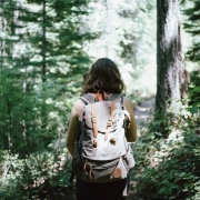 girl with brown hair walking through the woods with a backpack on